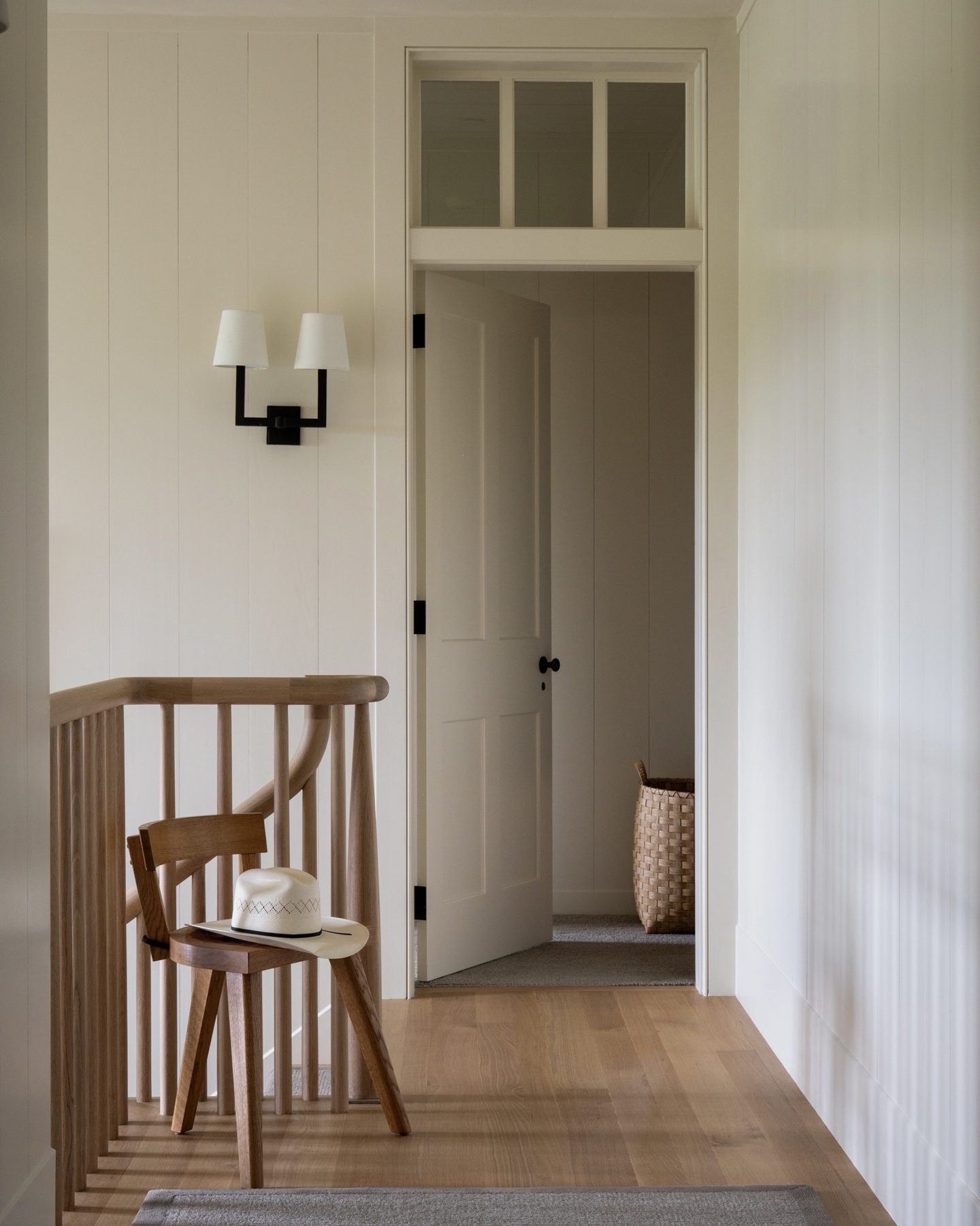 Stair landing with a wooden chair and straw hat, framed by an oak spindle railing and a paneled door beyond.