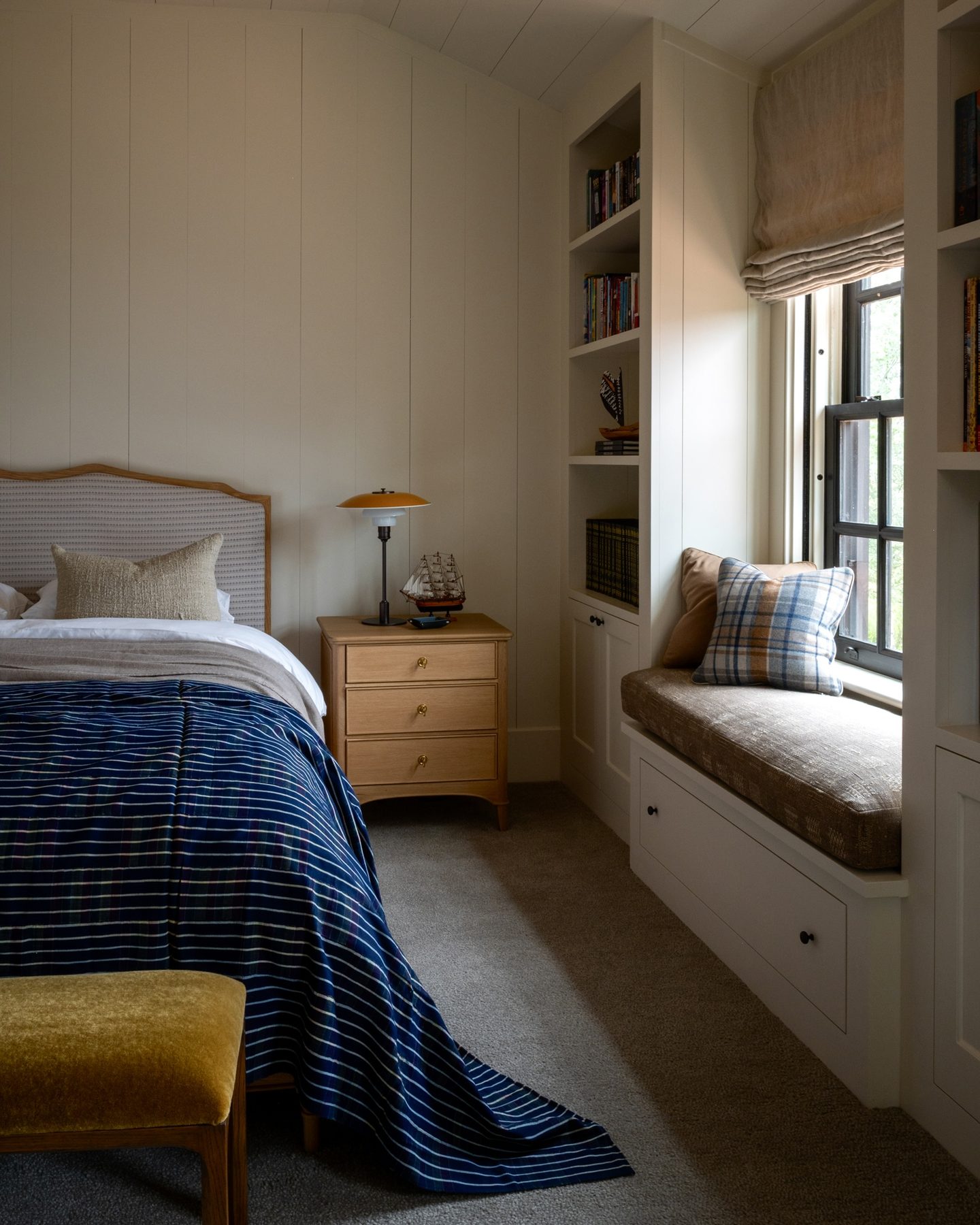 Bedroom with a striped indigo coverlet, a toy sailing ship on the nightstand, and a window seat layered with plaid and wool pillows.