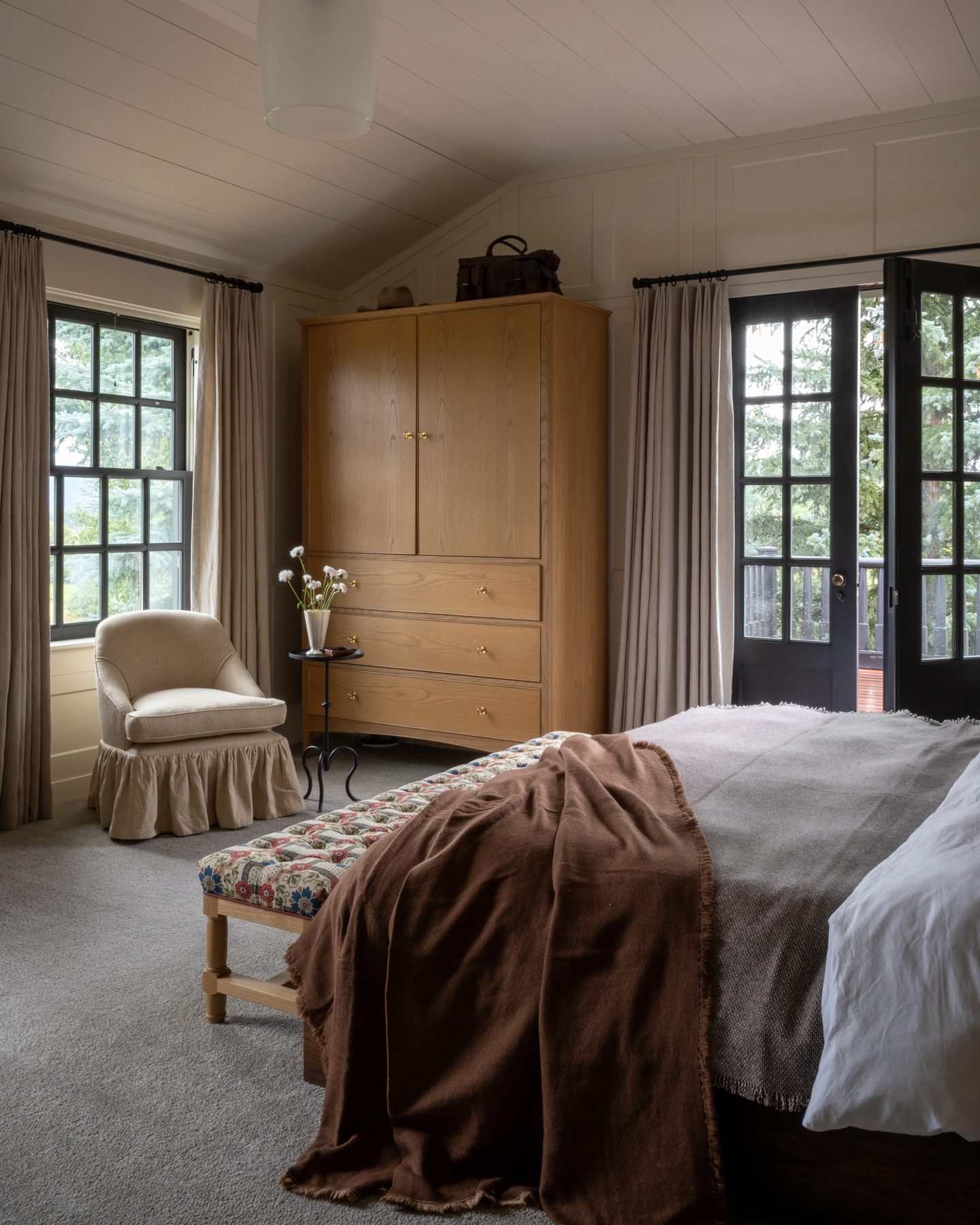 Bedroom corner with a paneled oak armoire, a skirted slipper chair, and a floral tufted bench draped in a wool throw.