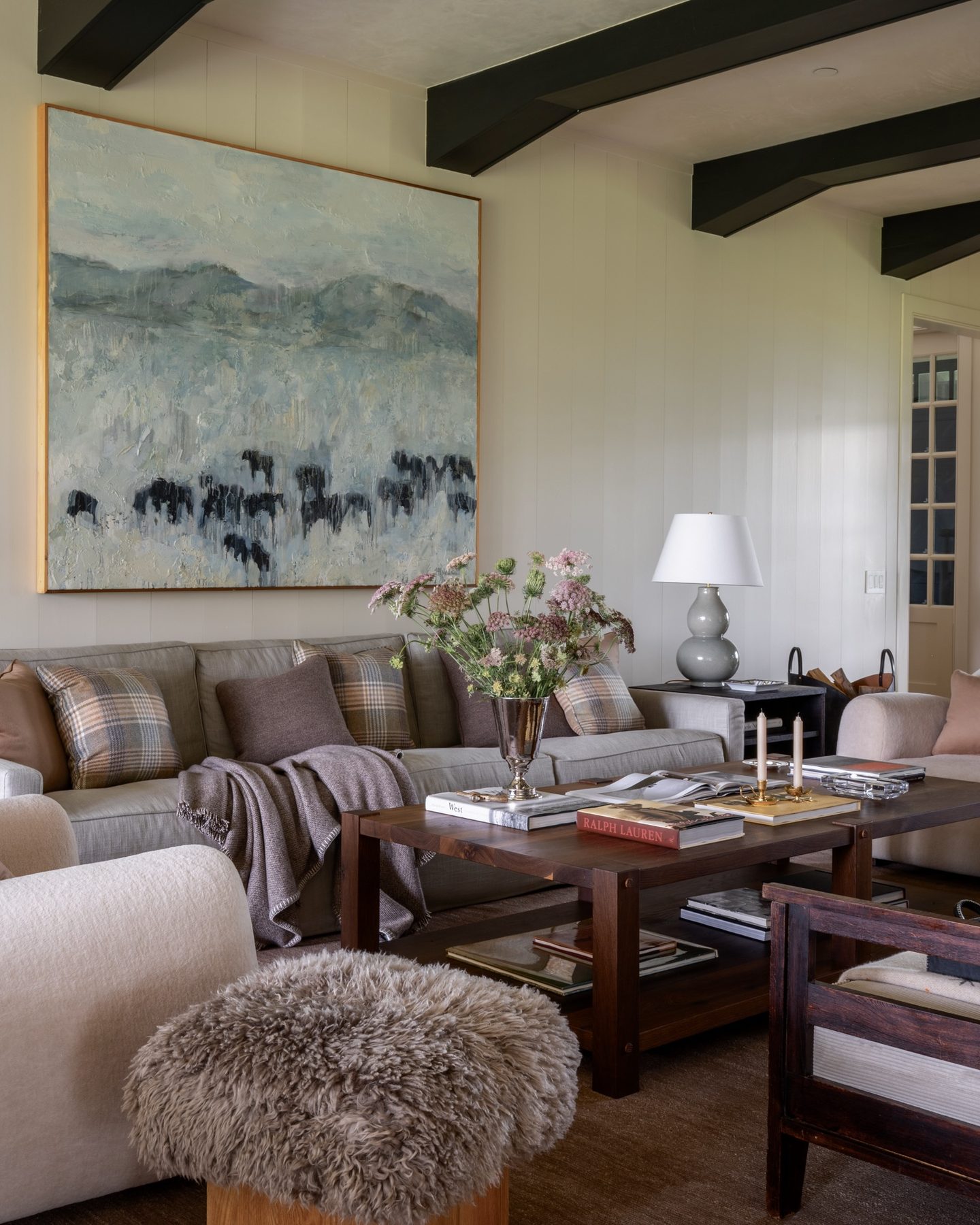 Living room with a horse-herd abstract painting above the sofa, plaid and mohair pillows, a sheepskin ottoman, and a stack of design books on the coffee table.