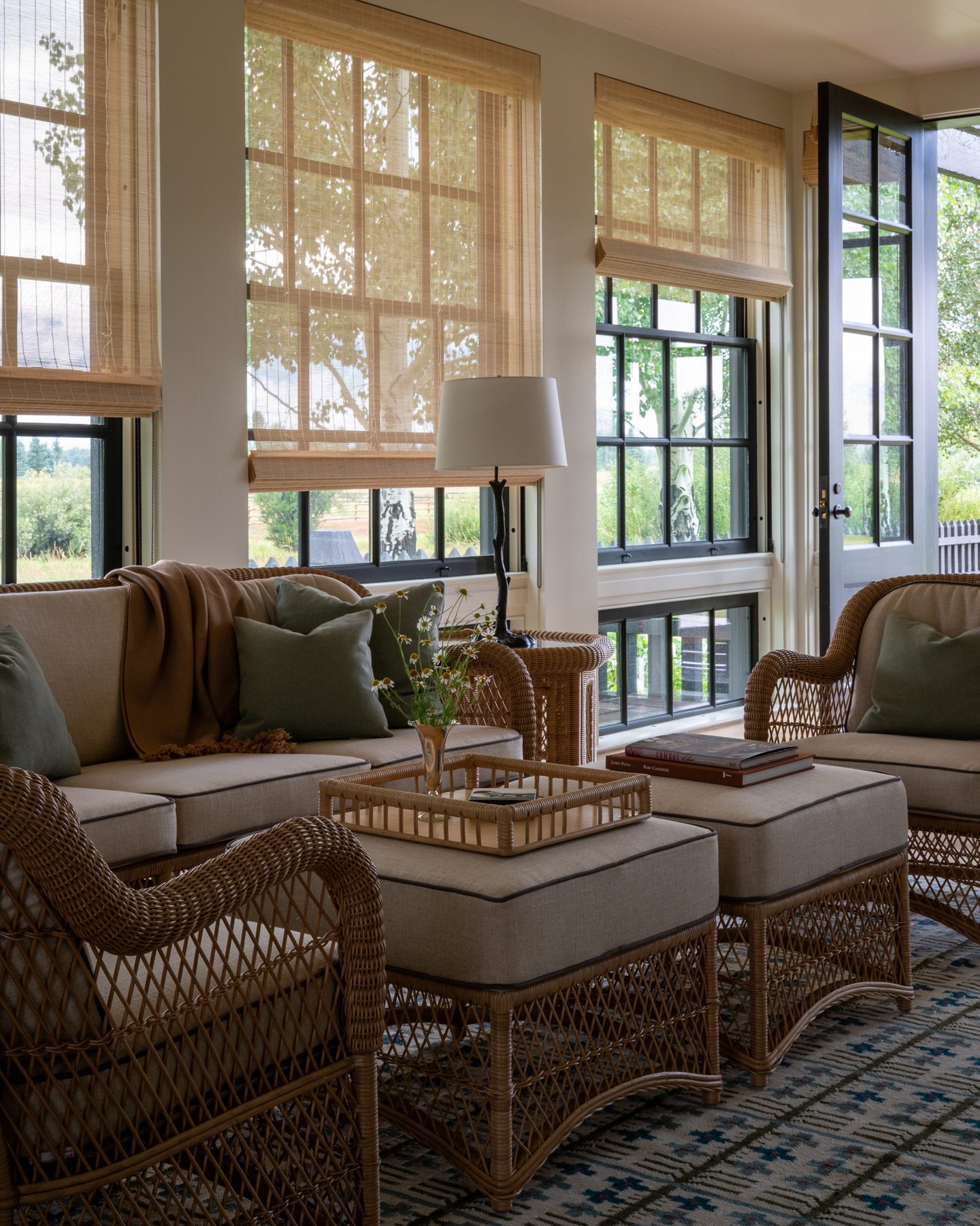 Screened sunroom with wicker armchairs, bamboo roman shades, and French doors opening to the meadow beyond.