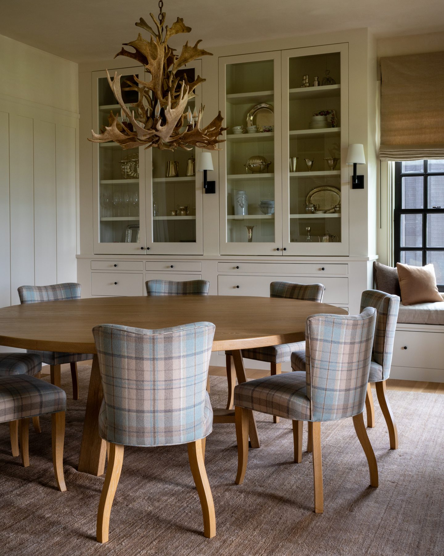 Dining room with a round oak table, plaid upholstered chairs, and glass-front cabinets beneath an antler chandelier.