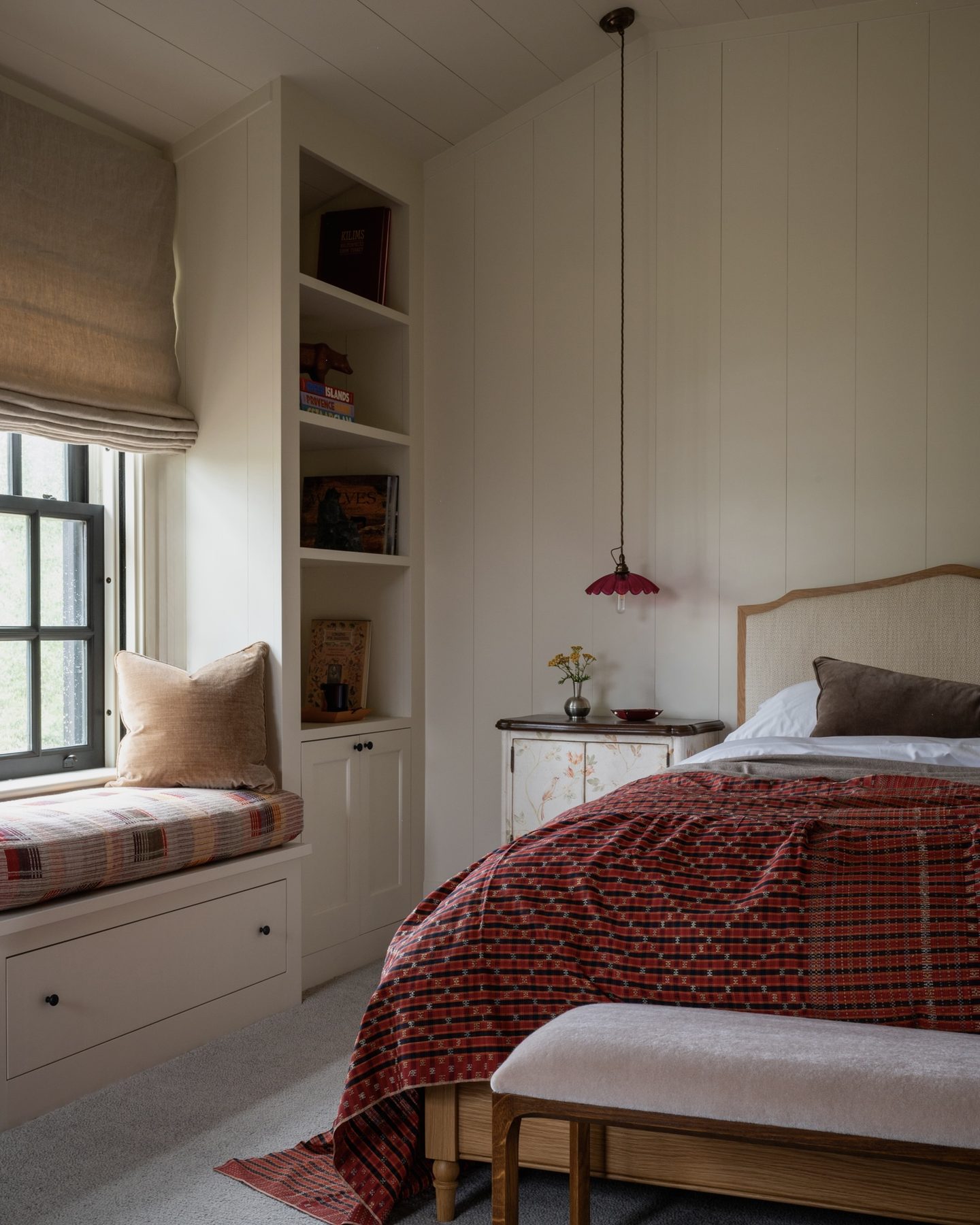 Bedroom with a caned headboard, red plaid coverlet, a window seat tucked beside built-in bookshelves, and a scalloped pink pendant light.