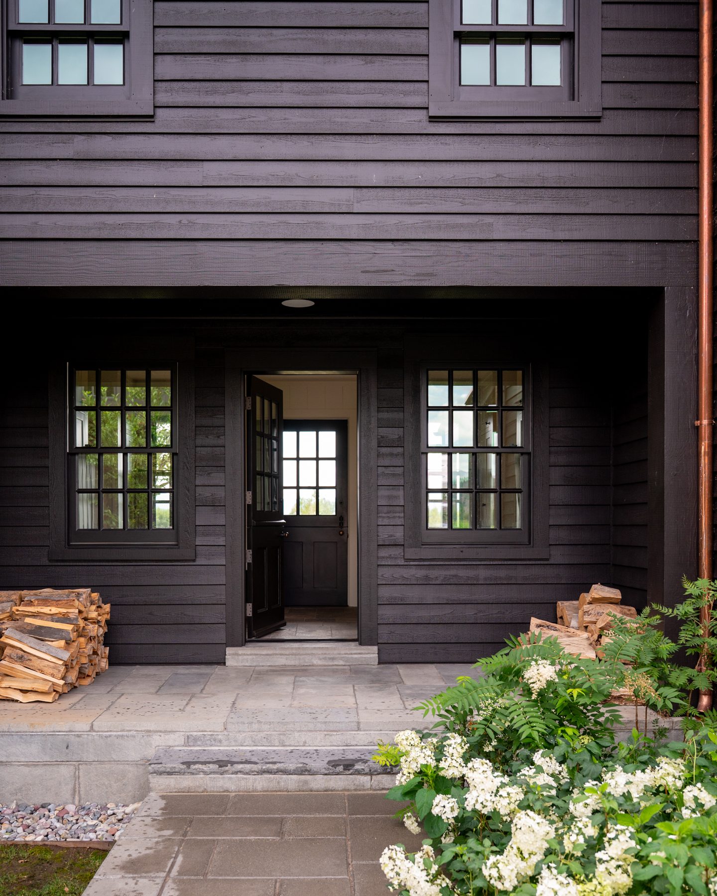 The residence's black timber facade with a Dutch door open to the entry, stacked firewood, and white hydrangeas in bloom along the front path.