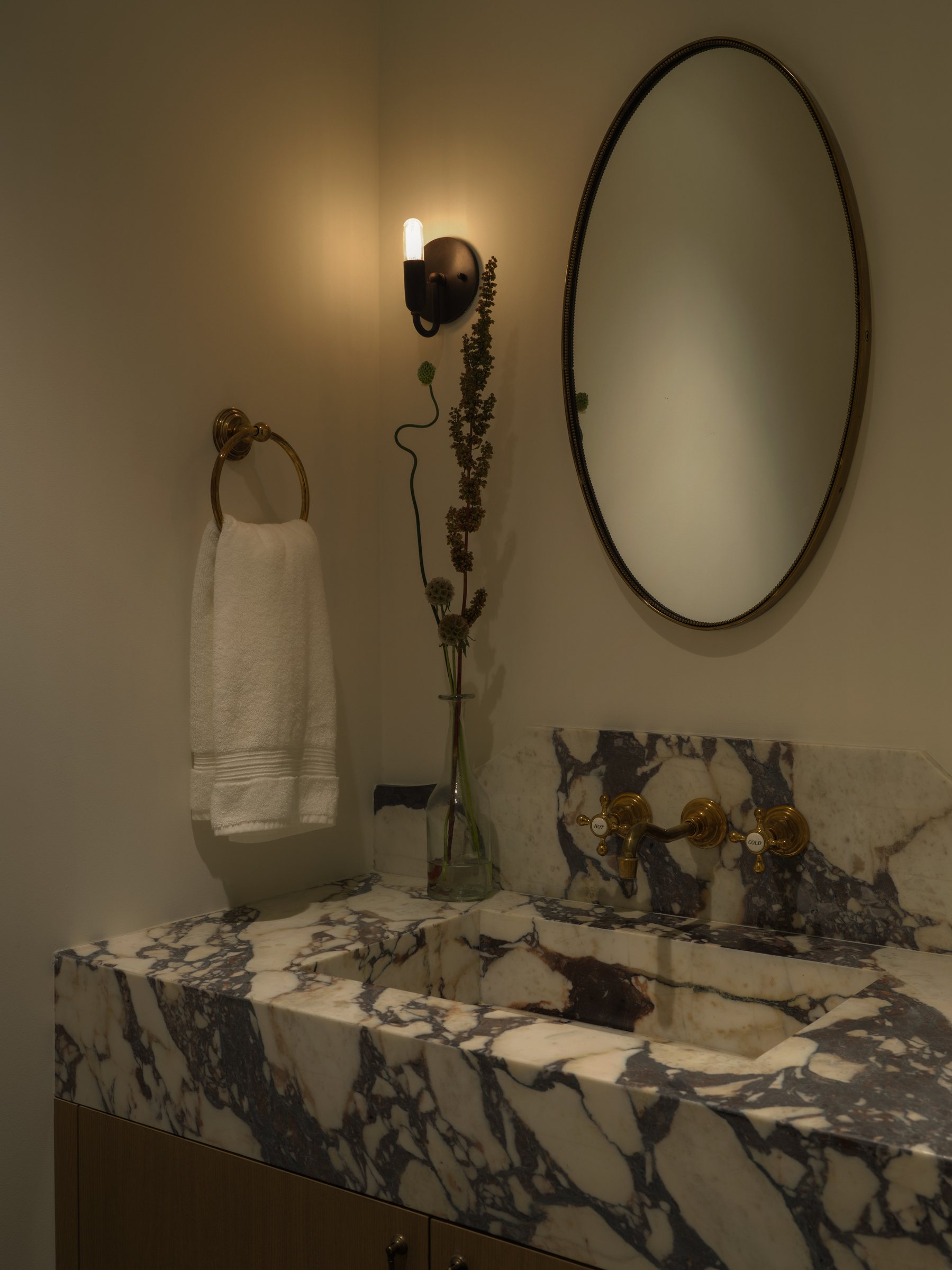 Marble vanity with brass fixtures, an oval mirror, and a wall sconce in the corner.