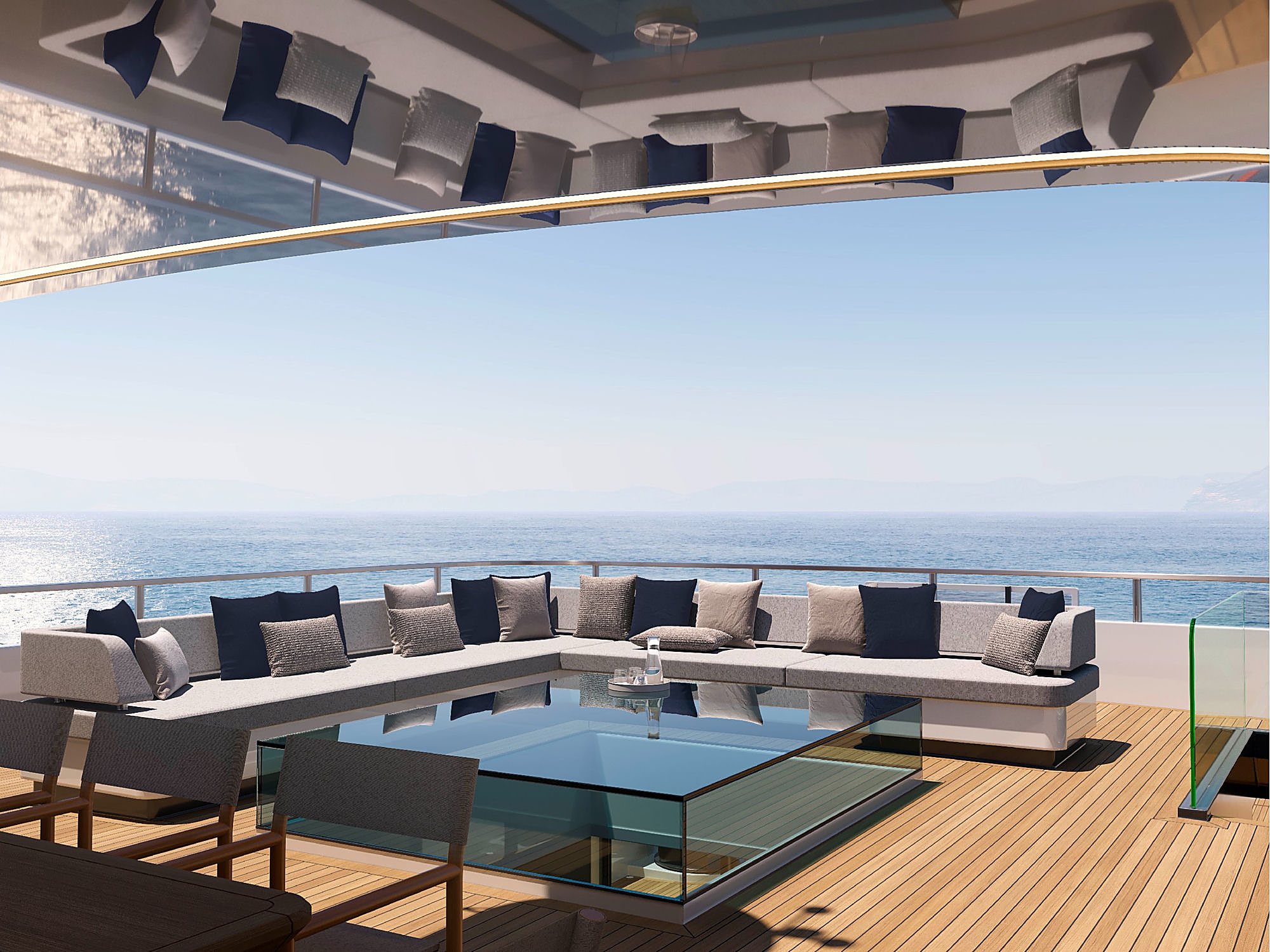 Aft deck lounge with a wraparound bench seat piled with navy and grey cushions, a glass-top coffee table, and open sea beyond the rail.
