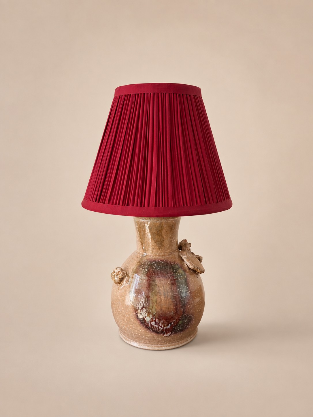 Ceramic table lamp by Jotham Hung with a mottled tan and oxblood glaze base, topped with a pleated crimson shade.