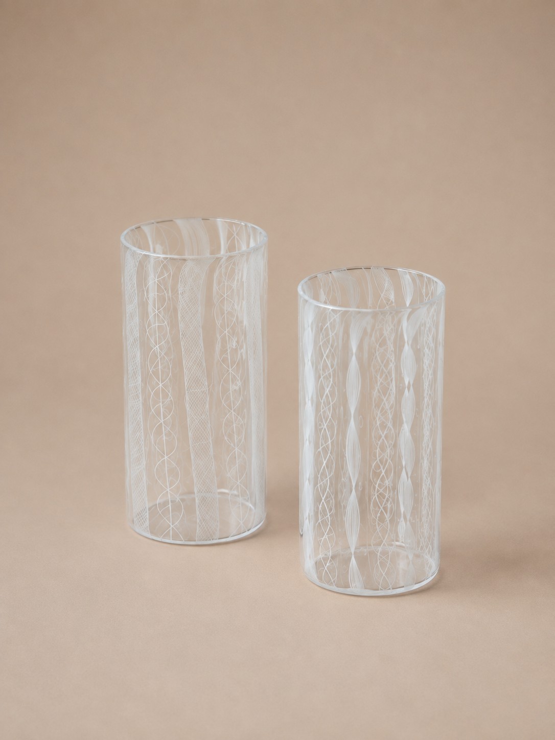 A pair of clear glass tumblers of two heights, each etched with fine vertical patterned bands, on a warm neutral background.