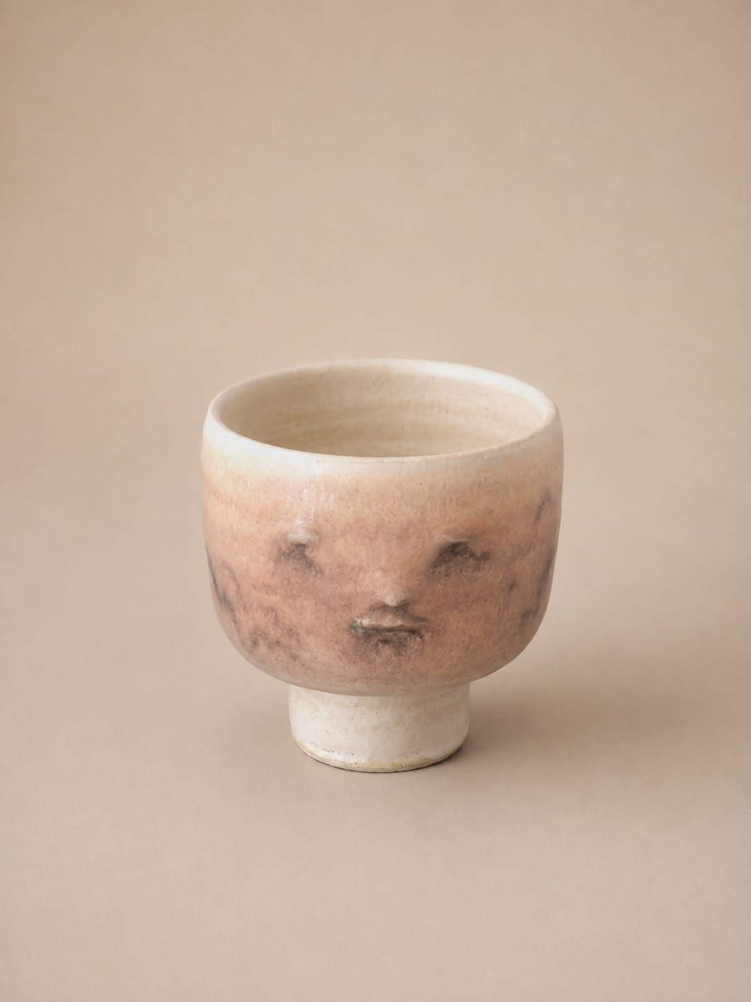 Ceramic footed cup by Cathrine Raben Davidsen with a sculpted face in a warm blush glaze, on a cream footed base.