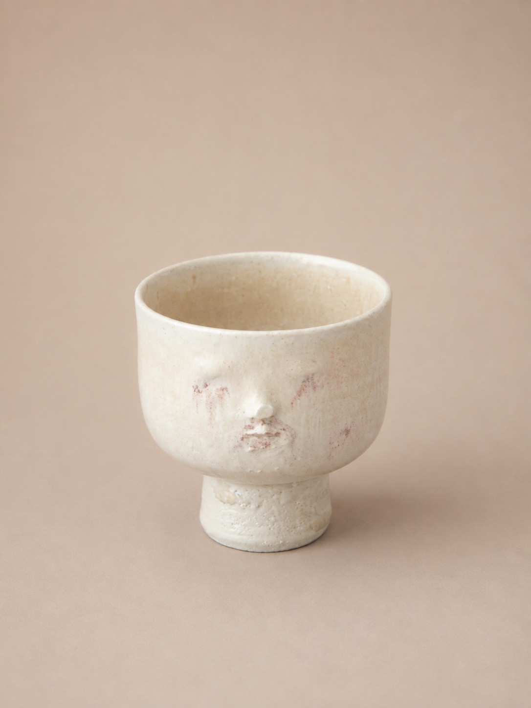 Ceramic footed cup by Cathrine Raben Davidsen with a sculpted face in a pale cream glaze with faint rose markings, on a matching footed base.