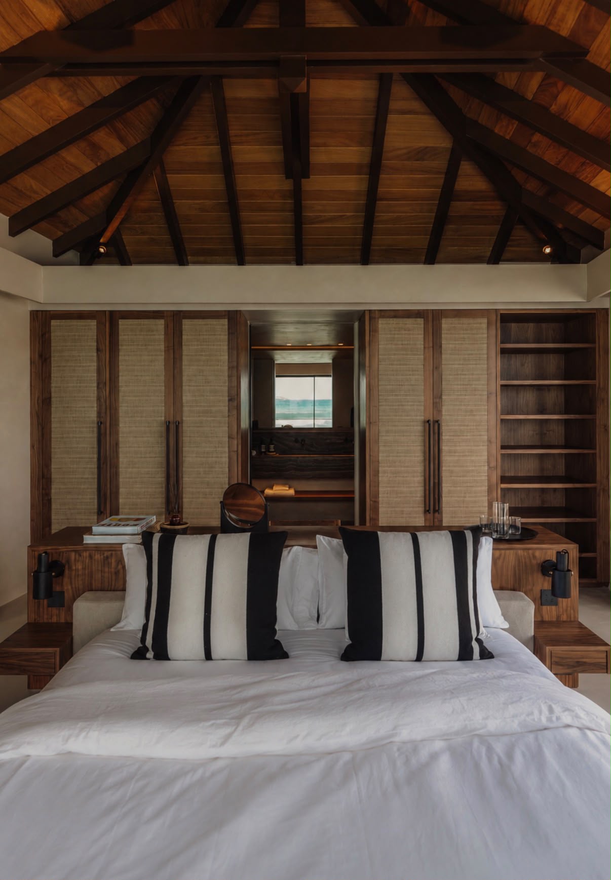 Bedroom with rattan-paneled closets, walnut nightstands, and a mirror reflecting the ensuite beyond.