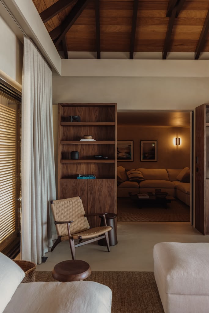 Reading nook with a woven lounge chair beside walnut shelving and linen curtains.