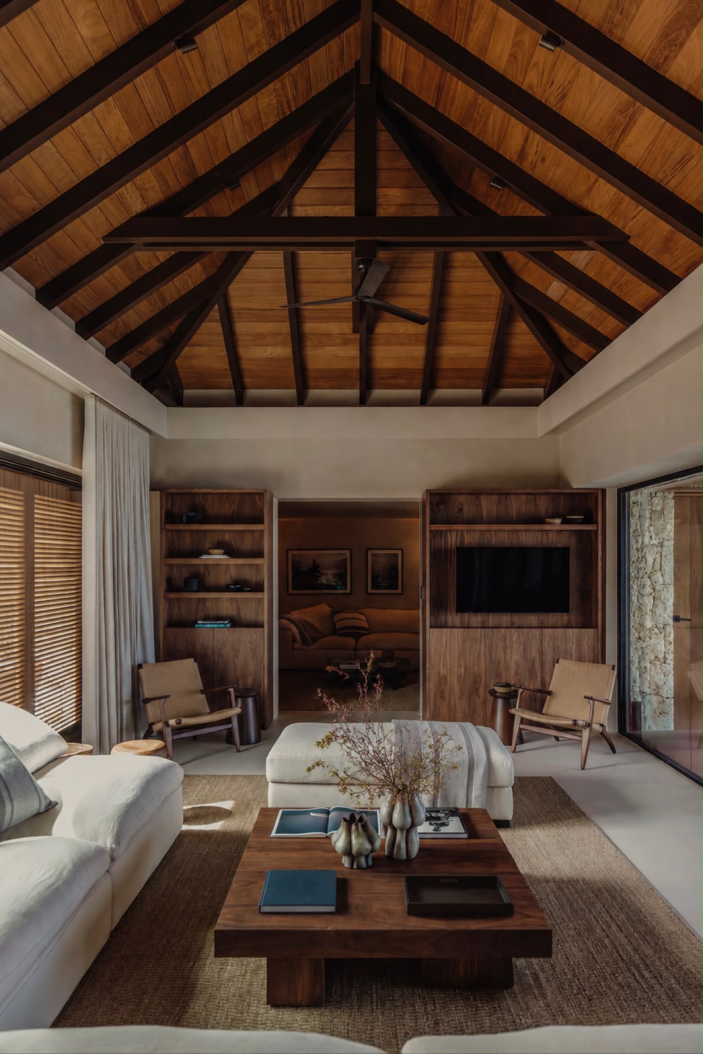 Living room with a vaulted wood-beamed ceiling, walnut built-ins, and woven lounge chairs around a linen ottoman.