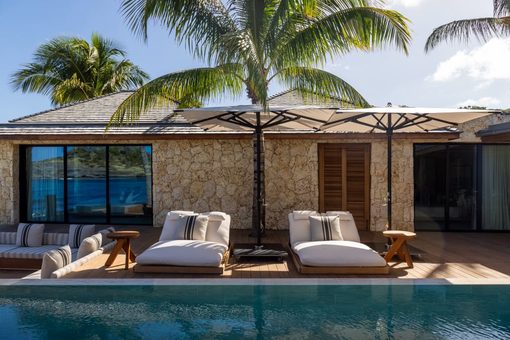 Pool deck with cushioned loungers, a shade umbrella, and the villa's stone facade beyond.